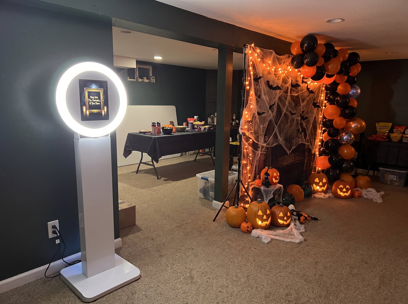The white photo booth showing a haunted portrait screen beside a Halloween backdrop of black and orange balloons, cobwebs and jack-o-lanterns