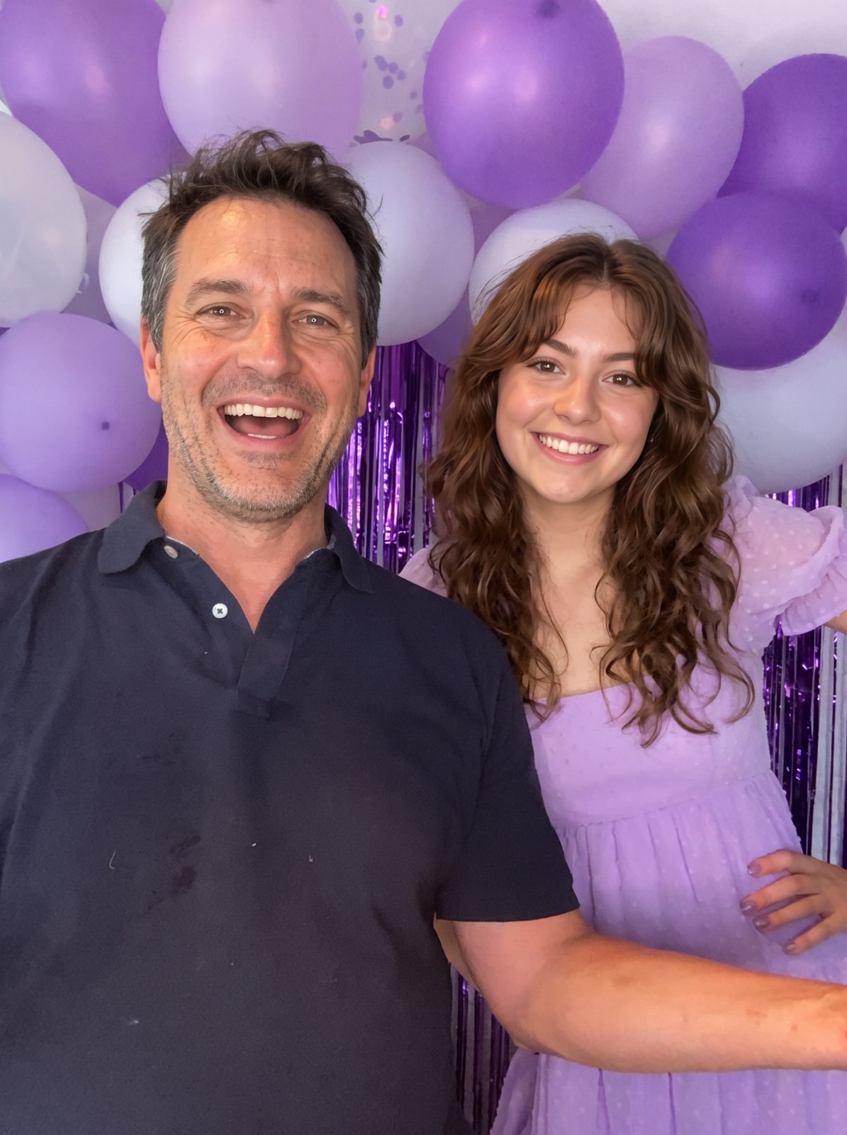 A laughing man and a smiling young woman in front of a purple and white balloon wall